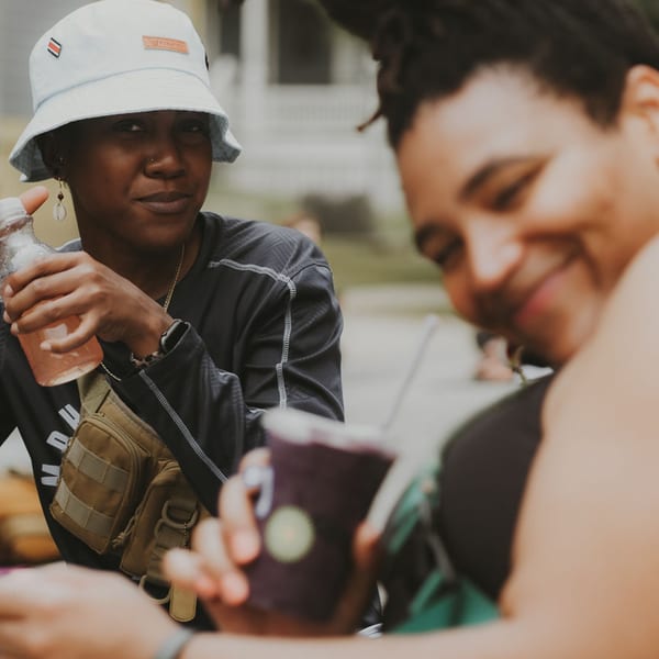 Two people enjoying drinks, one holding a bottle, the other a cup, in a casual outdoor setting.