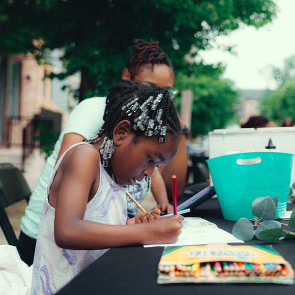 A young girl with beaded braids coloring at a table with an adult in the background.