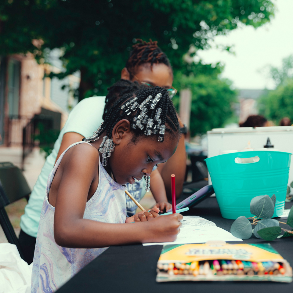 A young girl with beaded braids coloring at a table with an adult in the background.