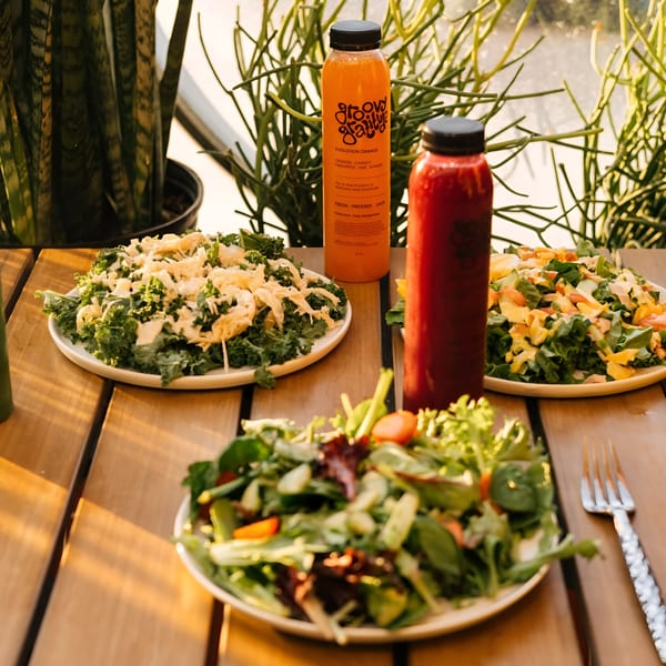 Three vibrant salads and two Groovy Gratitude juices on a sunlit wooden table.