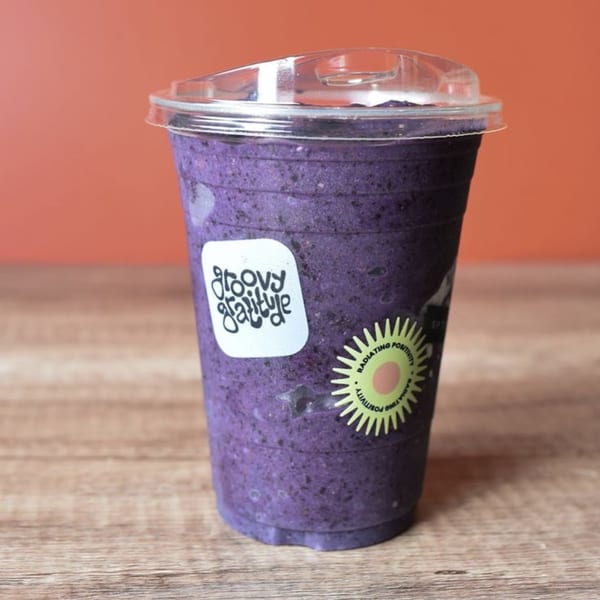 A Groovy Gratitude blueberry smoothie in a clear cup with a lid on a wooden table.