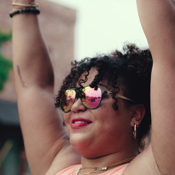 A woman with curly hair and pink sunglasses smiles with her arms raised.