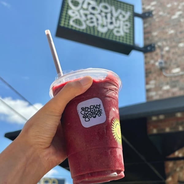 Hand holding a Groovy Gratitude berry smoothie with a straw, against a blue sky and restaurant sign.