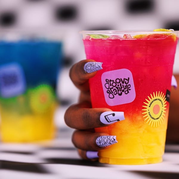 A hand with purple and black patterned nails holds a Groovy Gratitude pink and yellow drink.