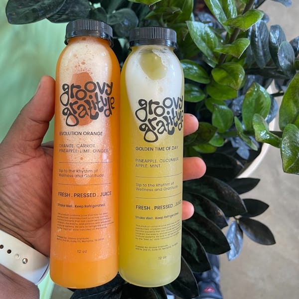 Two Groovy Gratitude juice bottles, orange "Evolution Orange" and yellow "Golden Time of Day", held against a plant.