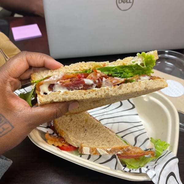 A hand holds a Groovy Gratitude BLT sandwich with bacon, lettuce, tomato, and mayo on whole wheat.