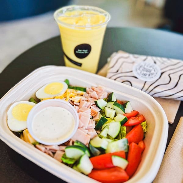A healthy Groovy Gratitude salad with ham, eggs, and vegetables, alongside a yellow smoothie.