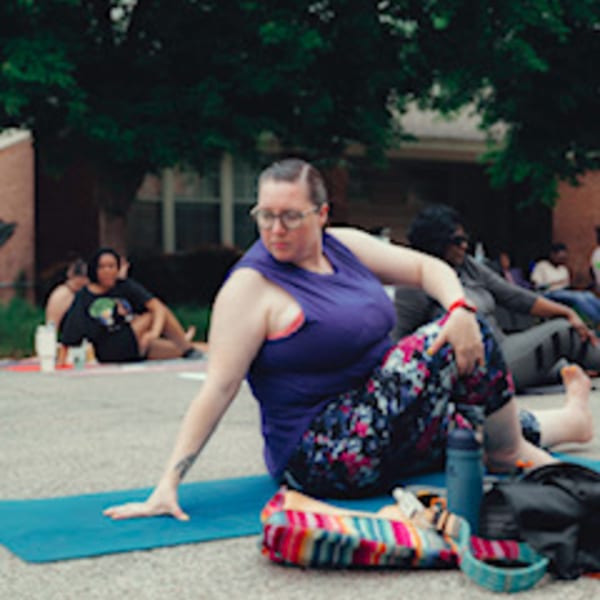 A woman in glasses and a purple top stretches on a blue yoga mat outdoors with others.